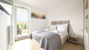 A bedroom with a bed, a nightstand, a small table, a painting, and a view of the outdoors.