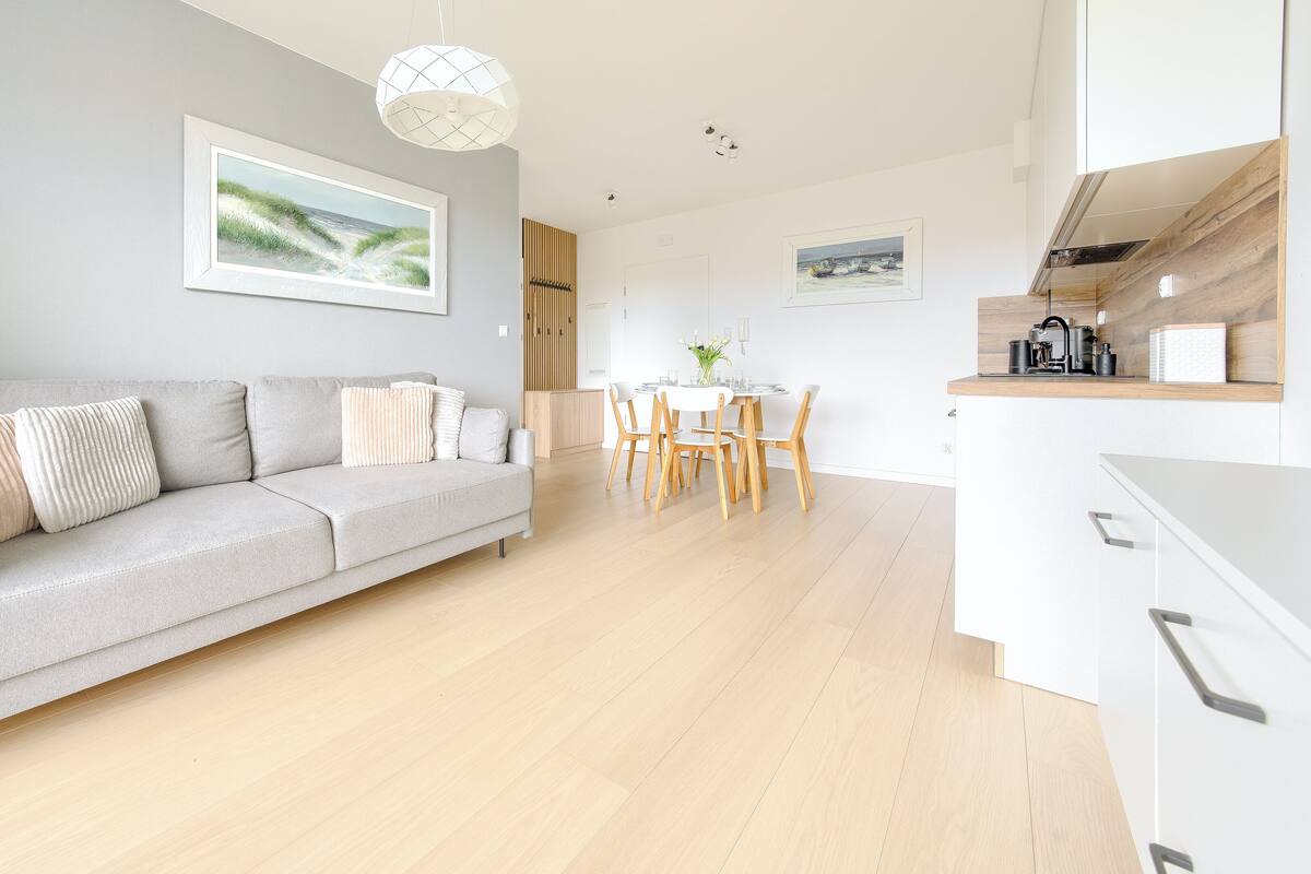 Una sala moderna con un sofá gris, mesa de comedor de madera y una cocina con armarios blancos.