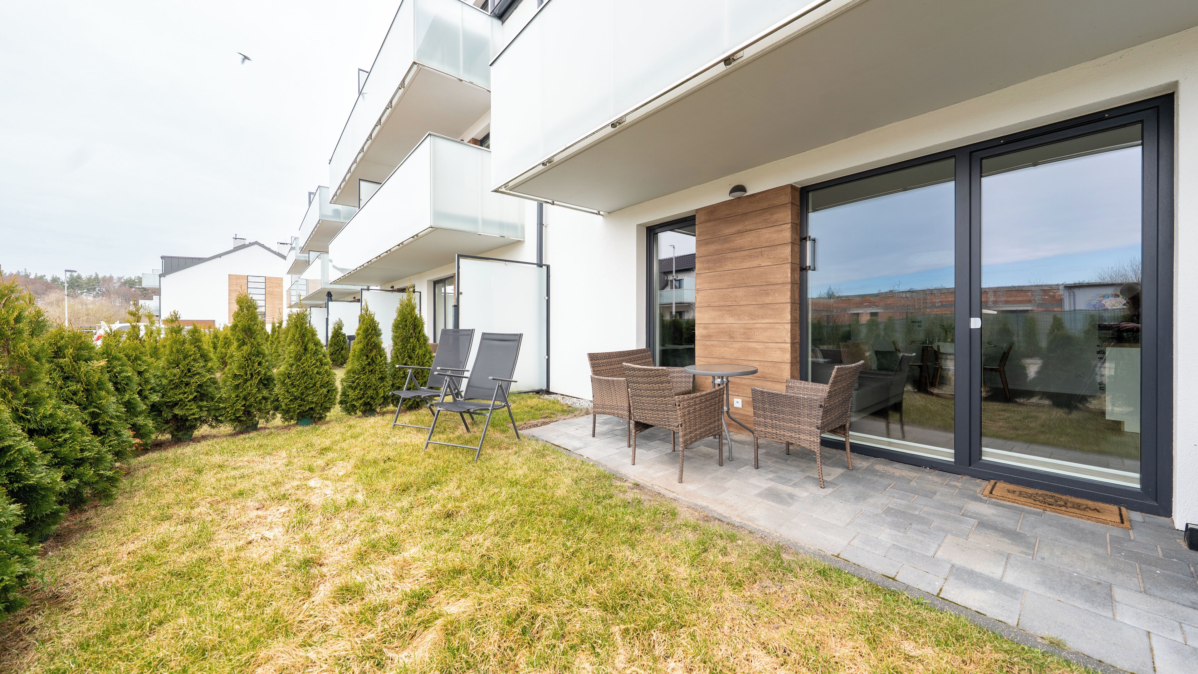 A modern outdoor patio with a seating area, a glass door leading to a garden, and a row of evergreen trees.