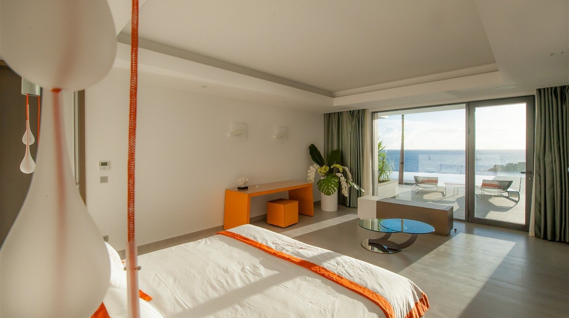 A modern hotel room with a large bed, a small desk, a glass table, and a balcony with a view of the sea.