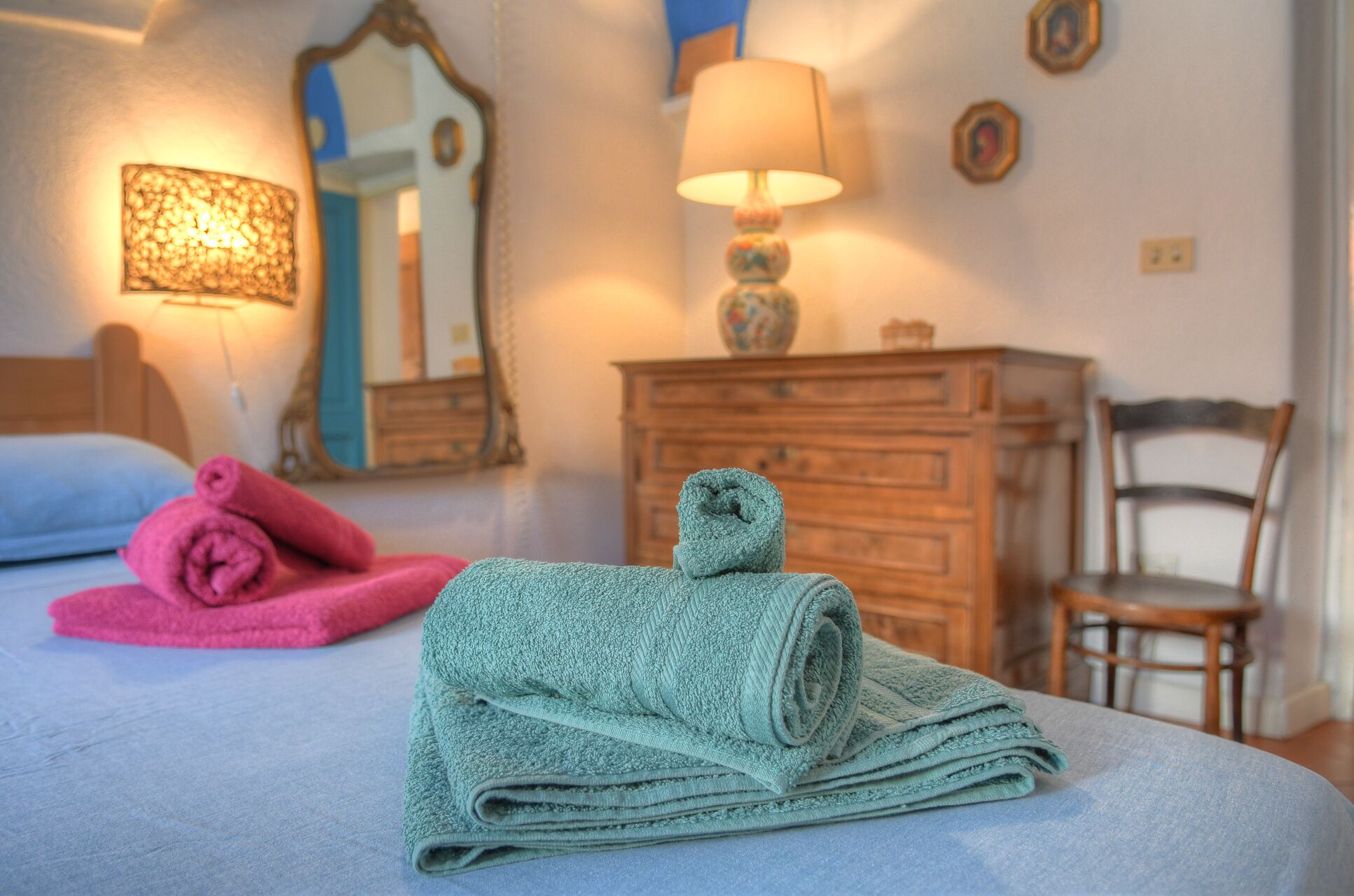 A neatly made bed with rolled towels, a wooden dresser, a chair, and a lamp.