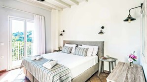 Un dormitorio con una cama grande, una mesita de noche, una pequeña mesa y una barra de noche. Se ve una ventana con cortinas, vista a zonas verdes y un techo con vigas a la vista.