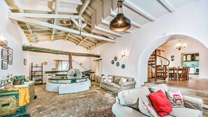 Amplia sala de estar con techo alto, chimenea de piedra en el centro, sofá con cojines decorativos y un área de comedor visible a través de un arco.