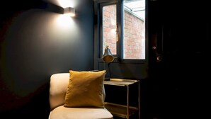 Una silla con cojín, una mesita con lámpara y una ventana con vista a un muro de ladrillo.