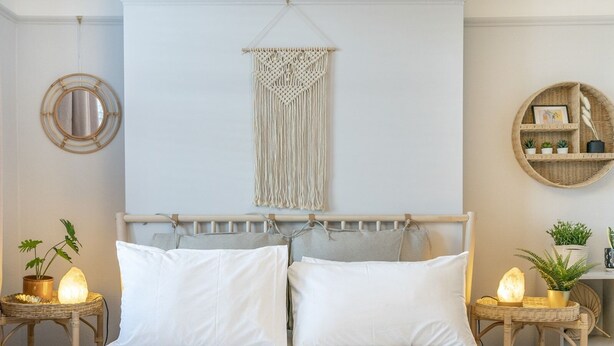 Una cama bien hecha con sábanas blancas, un colgador de macramé en la pared y dos mesitas de noche con lámparas y plantas.