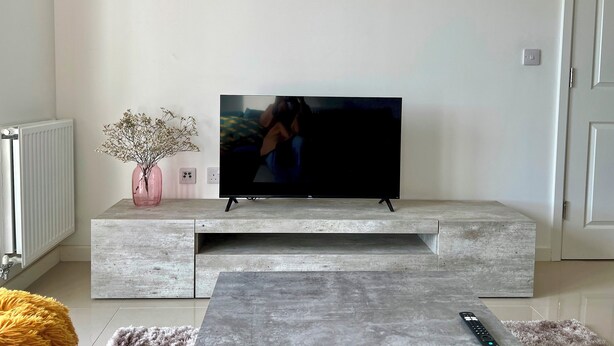 A living room with a flat-screen TV on a built-in shelf, a pink vase with dried flowers, a fluffy yellow bean bag, and a remote control on a gray rug. - Sleek 1 Bed Apartment w Parking - Pass the Keys (Cardiff)