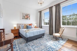 A bedroom with a large bed, two bedside tables, a chair, a chandelier, and a window with curtains. - Devonshire House Luxury with a Fascinating History (North Hobart)