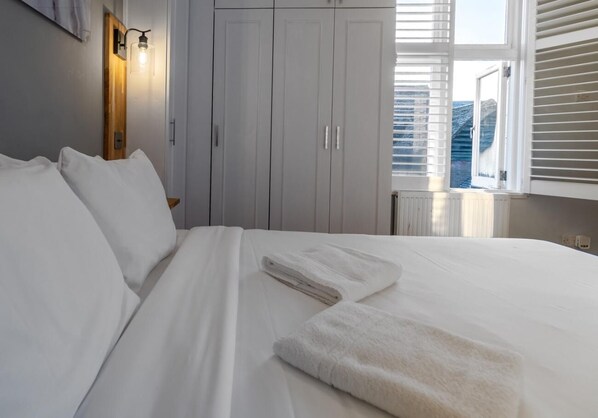 Una cama bien tendida con sábanas blancas y una toalla doblada, una mesita de noche de madera con una lámpara, y un amplio armario con puertas correderas.