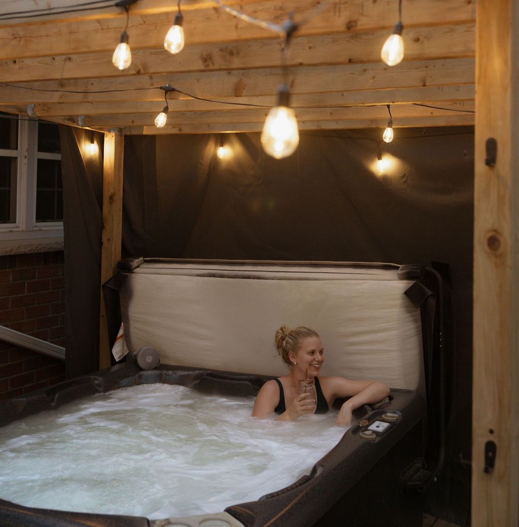 Un bain à remous avec une personne à l’intérieur, entouré de murs en bois et illuminé de guirlandes lumineuses.