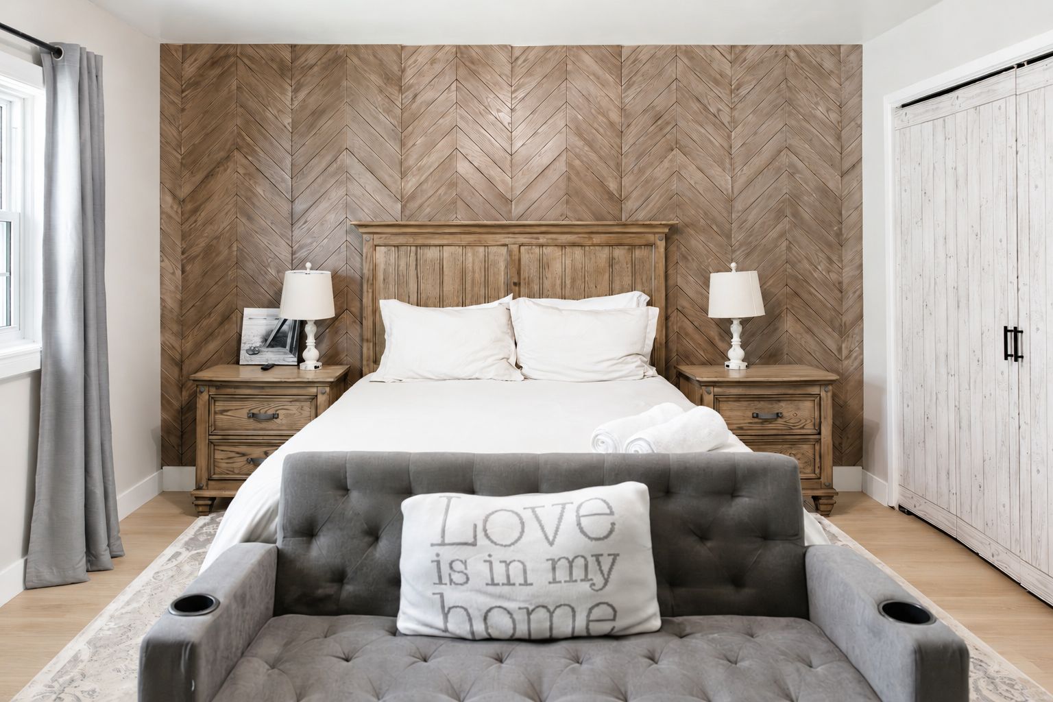 A bedroom with a herringbone-patterned wall, a wooden headboard, a grey upholstered armchair, and a pillow with 'Love is in my home' text.