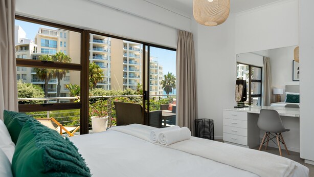 Une chambre d’hôtel avec un grand lit, un bureau et un balcon offrant une vue sur d’autres immeubles et sur la verdure.