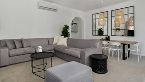 Un salon moderne avec un canapé d’angle gris, une table basse ronde et un coin repas comprenant une table en bois et des chaises blanches.