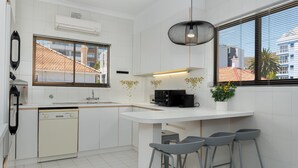 Une cuisine moderne avec des meubles blancs, un lave-vaisselle encastré, une machine à café noire et une fenêtre donnant sur des immeubles. On y trouve trois tabourets de bar gris et un luminaire suspendu.