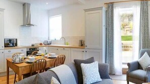A modern kitchen with white cabinetry, a dining area with a wooden table and chairs, and a grey sofa with a patterned throw.