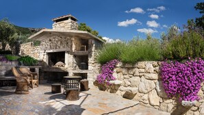 Un bâtiment en pierre doté d’une cheminée, d’un espace extérieur aménagé pour s’asseoir et de fleurs violettes.