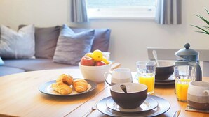 Une table en bois dressée pour le petit-déjeuner, avec des croissants, des fruits, du café et du jus d’orange.