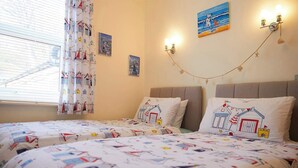 Une chambre avec deux lits, de la literie sur le thème de la marine, une fenêtre avec des rideaux, des œuvres d’art murales et une guirlande lumineuse.