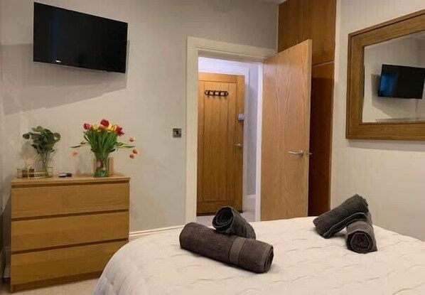 A hotel room with a bed, a wooden dresser, a flat-screen TV mounted on the wall, and a door leading to another room.