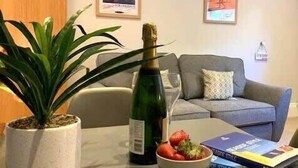 A living room with a sofa, a potted plant, a bottle of champagne, a bowl of strawberries, and a magazine on the coffee table.