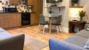 A compact living space with a kitchenette, a dining area with two chairs, a flat-screen TV mounted on the wall, and a small side table with a lamp and flowers.