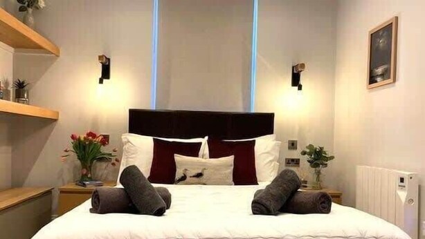 A neatly made bed with a white duvet, two pillows, and two rolled towels. There are two bedside tables with lamps, a wall-mounted shelf with decorative items, and a framed picture on the wall.