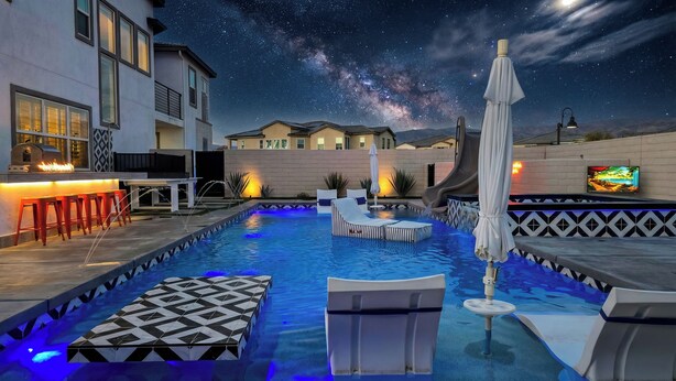 A swimming pool with lounge chairs and a bar area, surrounded by houses under a starry night sky. - Ultimate Desert Getaway, Pool/Spa, 4k Game Room (Indio)