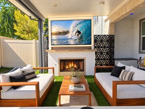 A modern outdoor living space with a fireplace, comfortable seating, and a large TV screen displaying a surfing scene. - Ultimate Desert Getaway, Pool/Spa, 4k Game Room (Indio)