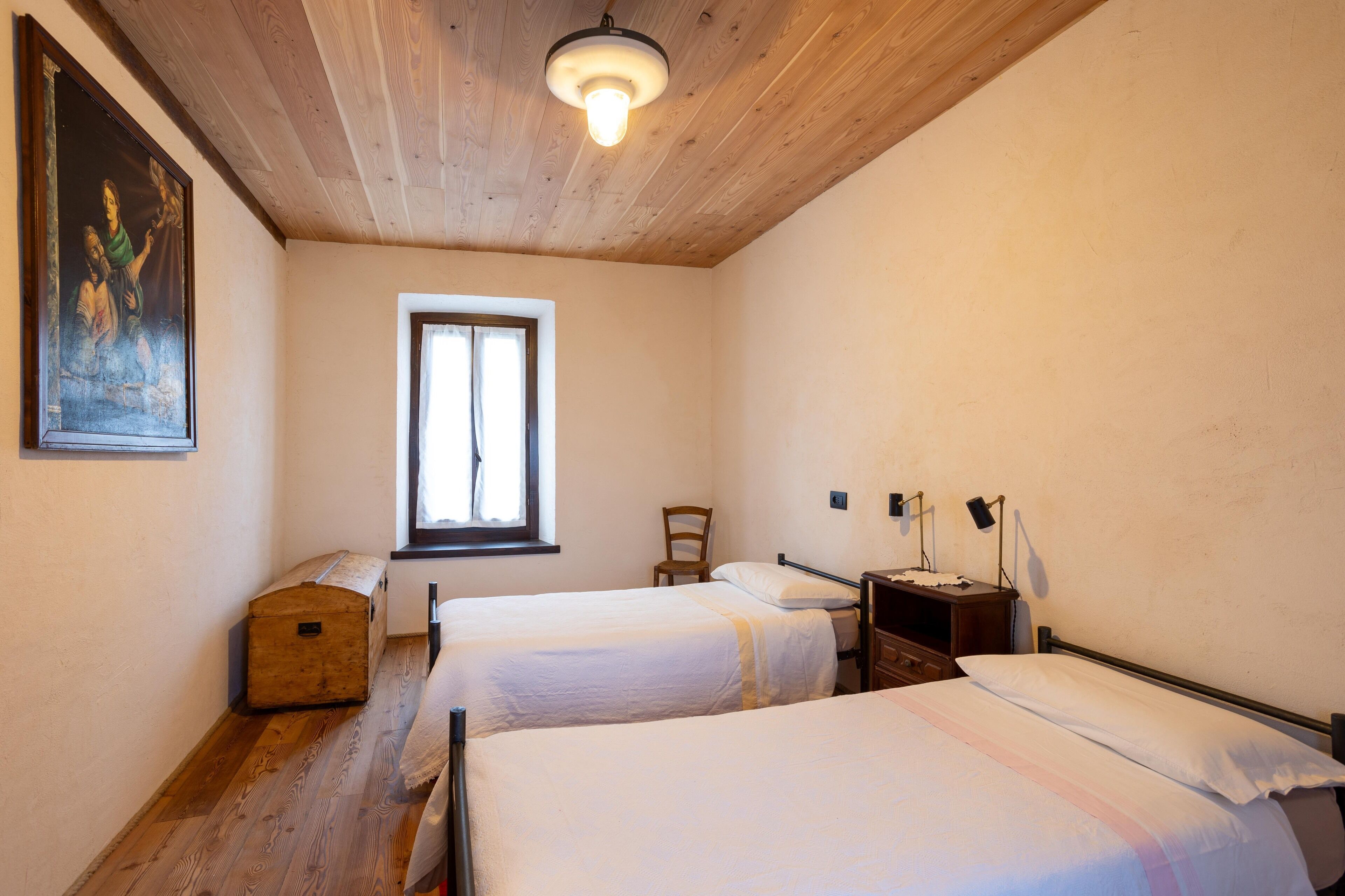 A room with two beds, a wooden ceiling, a window, a chair, and a painting on the wall.