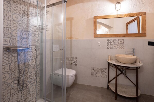 Casa de banho com azulejos estampados, cabine de chuveiro com vidro, sanita e lavatório com pedestal de madeira.
