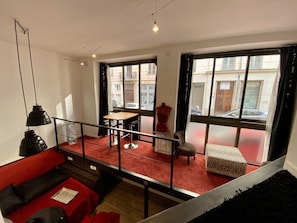 A modern apartment with a red carpet, a dining area, a sofa, and large windows offering a view of the street.