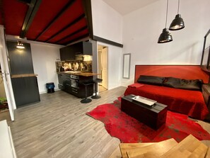 A compact hotel room with a kitchenette, a bed with red bedding, a red rug, and two pendant lights.
