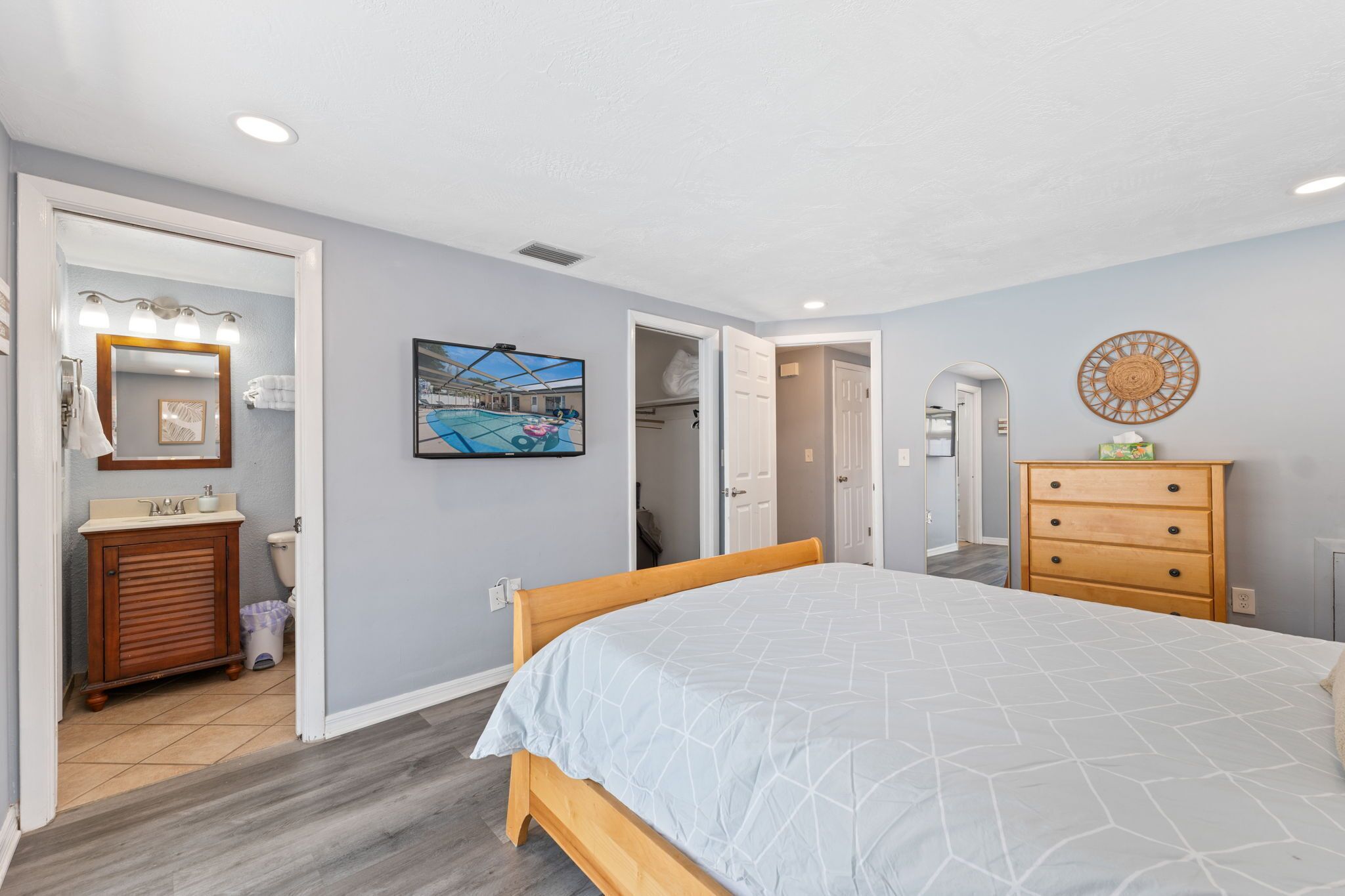 Un dormitorio con cama, cómoda de madera, baño con lavamanos y espejo, y un cuadro en la pared.