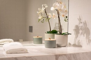 Une salle de bain avec des carreaux blancs, un comptoir en marbre, une serviette et trois plantes en pot ornées de fleurs blanches.