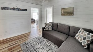 A room with a gray sofa, patterned rug, wooden flooring, white walls, and a ceiling fan. - Toes in the Sand | Pet-Friendly Downtown Cottage Steps from the Beach! (Carolina Beach)
