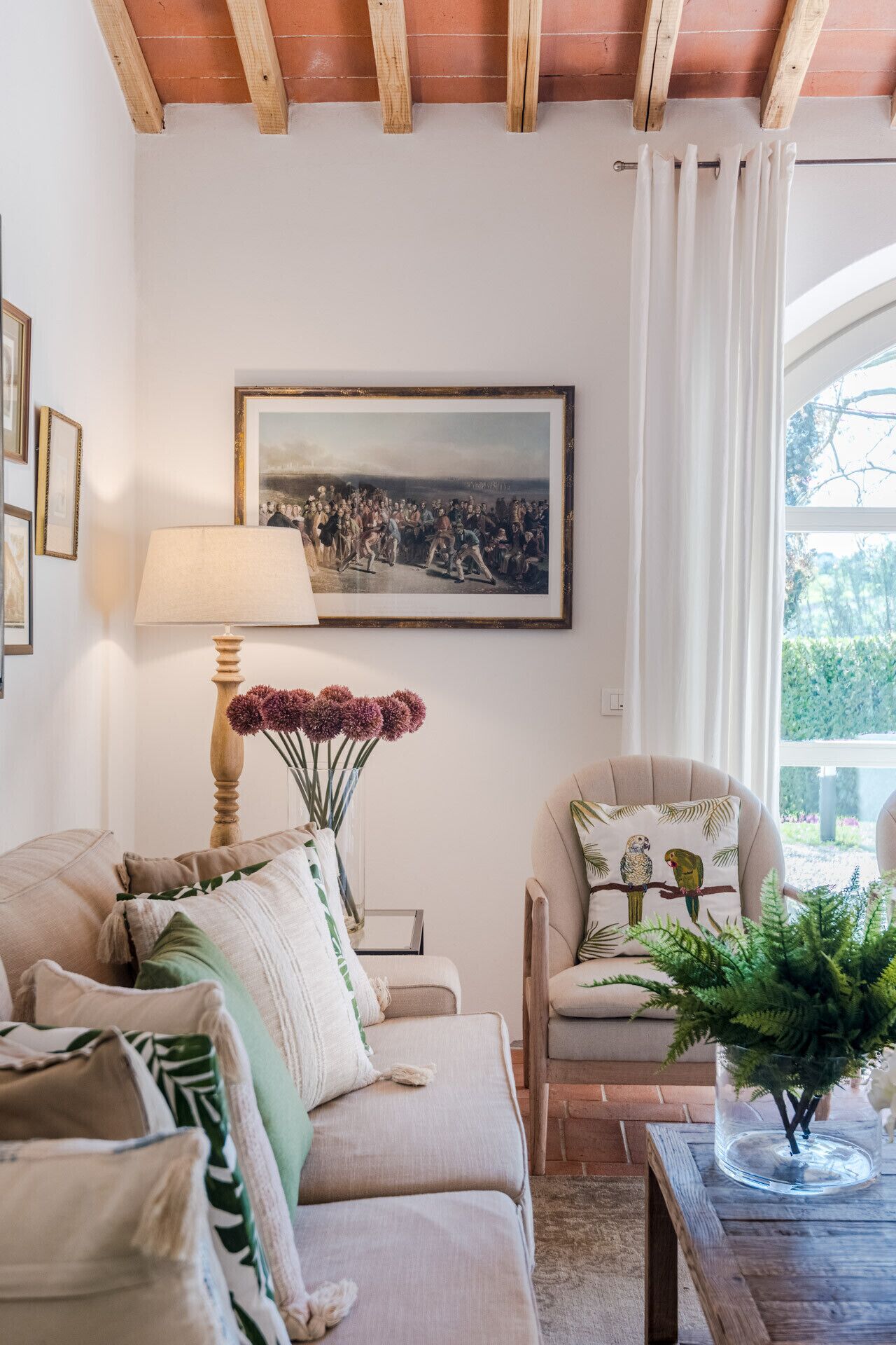 A cozy living room with a sofa, armchair, and a table with a vase. A painting hangs on the wall, and there's a window with curtains.