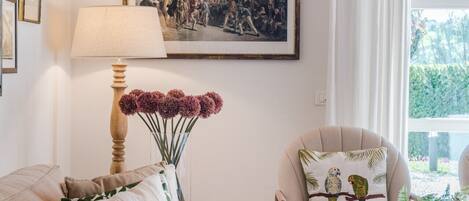 A cozy living room with a sofa, armchair, and a table with a vase. A painting hangs on the wall, and there's a window with curtains.