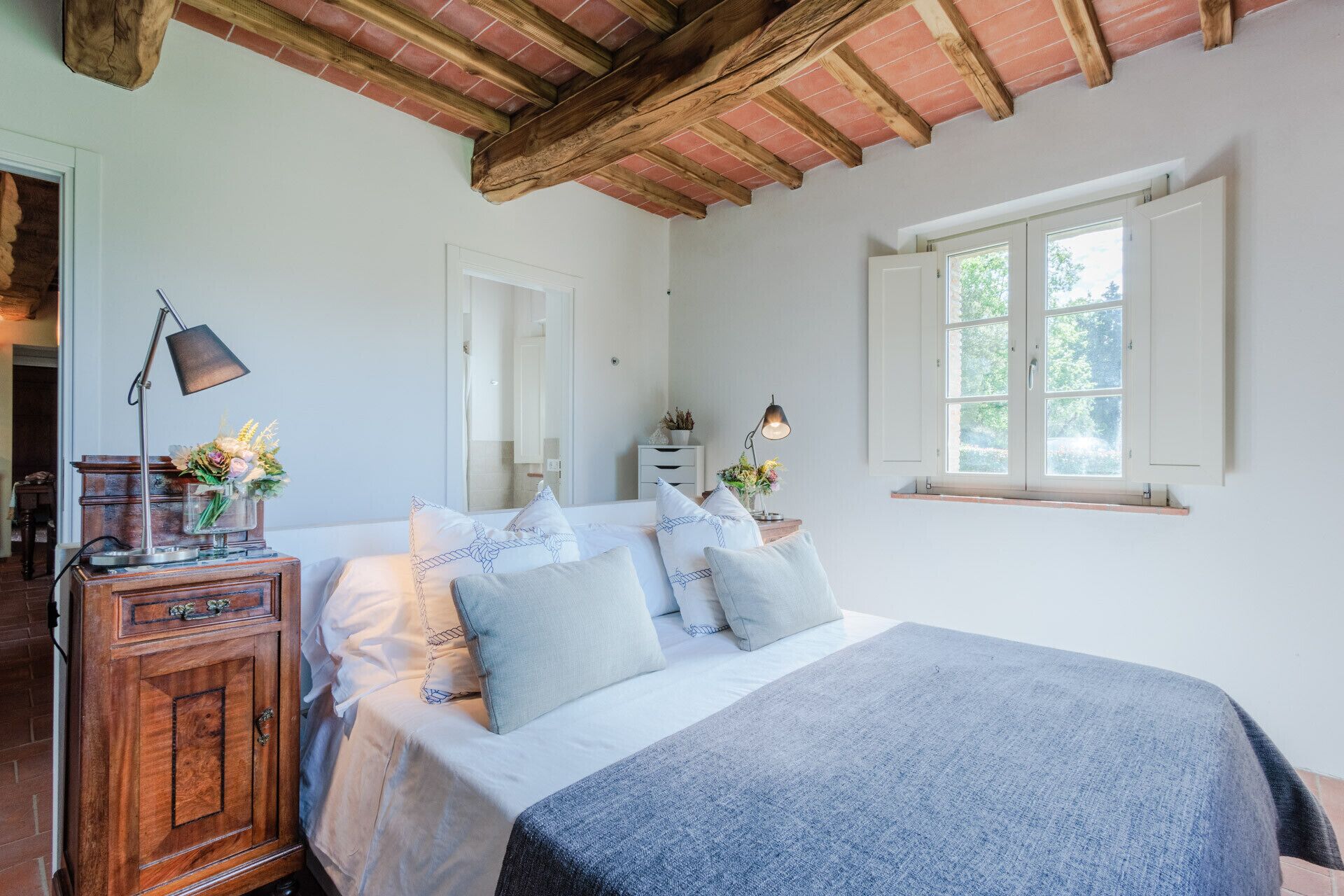 A bedroom with a wooden headboard, a large bed with multiple pillows, a nightstand with a lamp, a dresser, and a window with a view of greenery.