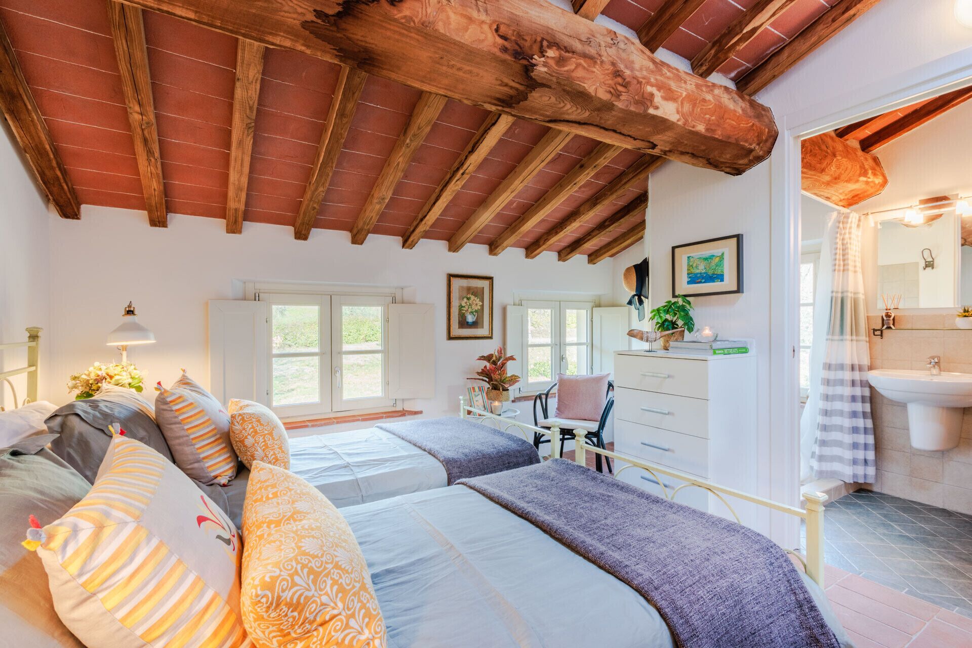 A bedroom with two beds, a wooden ceiling, a window, and a bathroom visible through an open door.