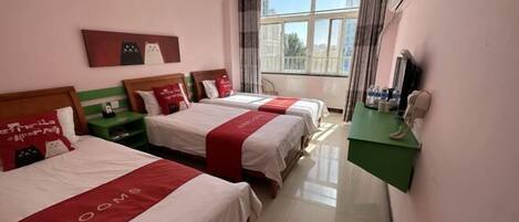 A hotel room with three beds, a green desk, a window with curtains, and a wall-mounted air conditioning unit.
