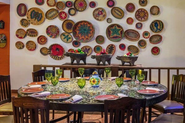 A dining area with a round table set for four, surrounded by chairs. The table is covered with a patterned tablecloth and set with plates, glasses, and a decorative vase.