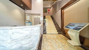 Un vestíbulo de hotel con un mostrador de recepción de mármol, un quiosco informativo digital, una escalera y un elemento decorativo rojo en la pared.