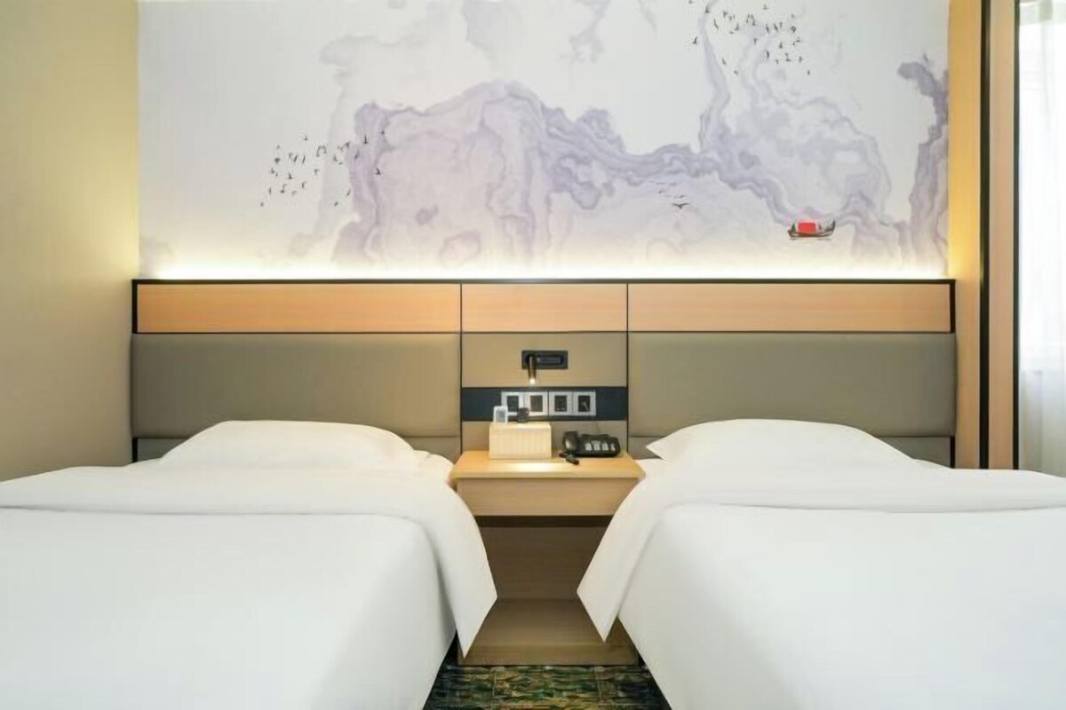 Une chambre d’hôtel avec deux lits, une table de chevet, un panneau de commande fixé au mur et une grande œuvre d’art abstraite.
