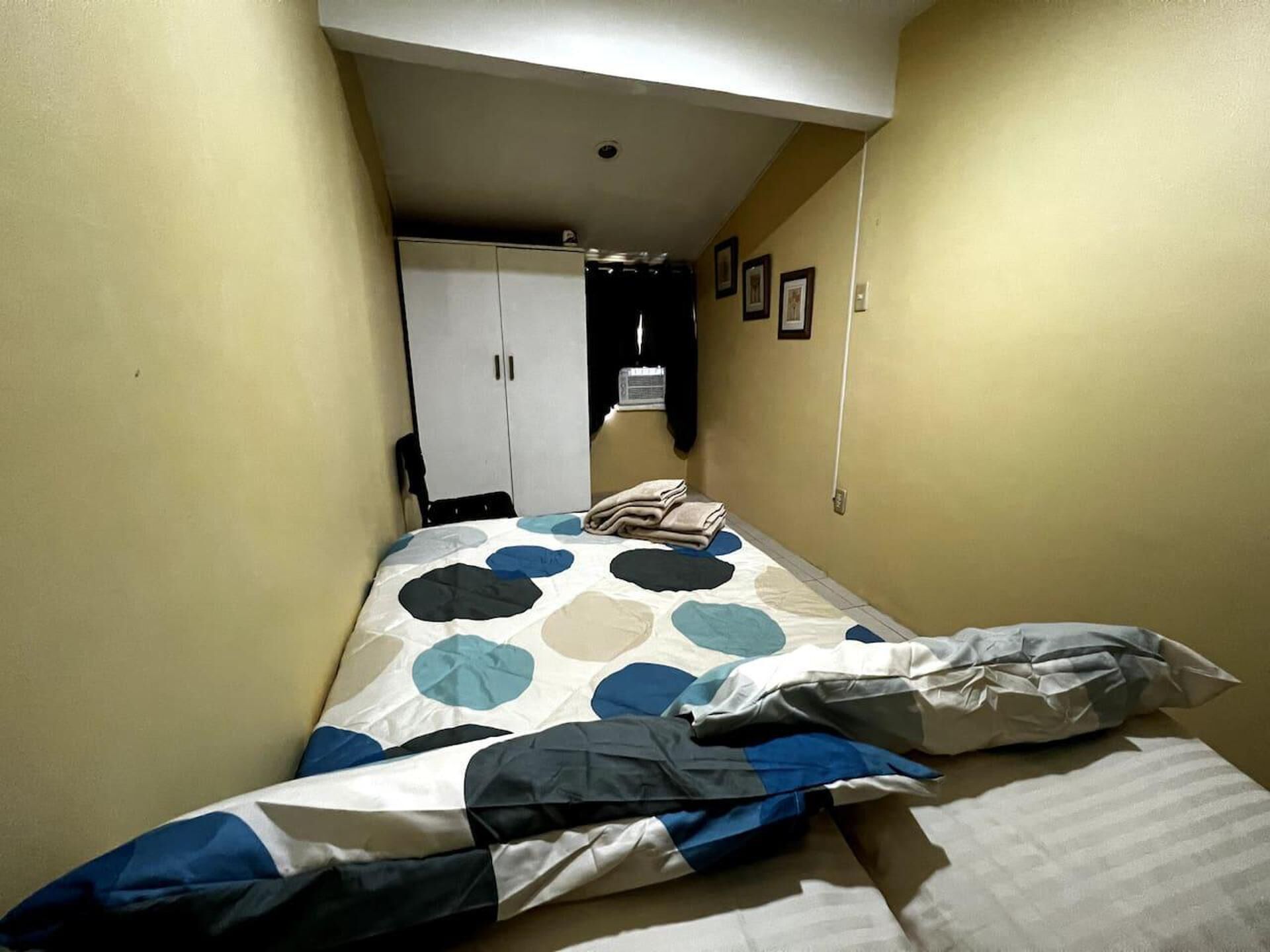 A small, narrow room with a bed, a wardrobe, and a door leading to another room.