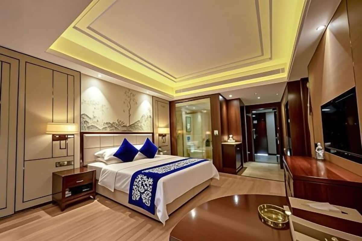 A modern hotel room with a bed, bedside tables, a TV, and a bathroom visible through a glass door.