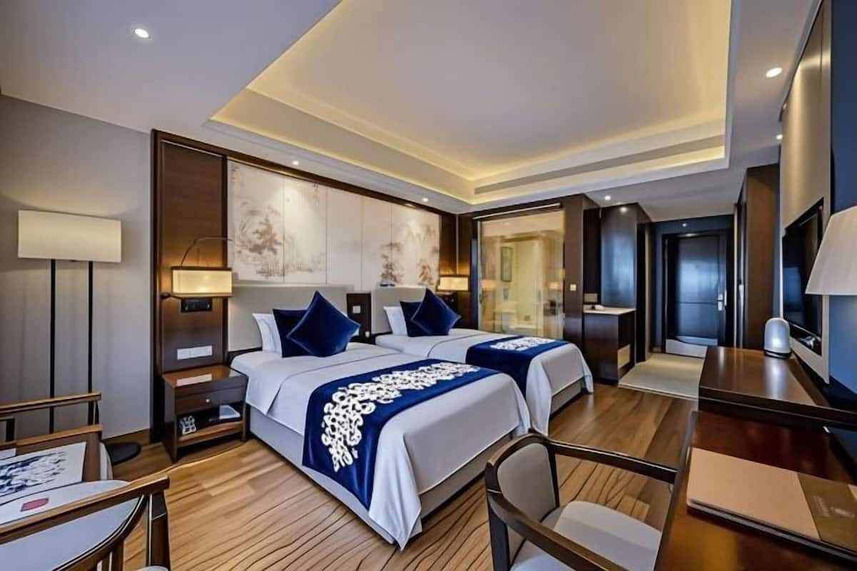 A hotel room with two beds, a desk, a chair, a TV, and artwork on the walls.