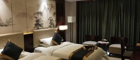 A hotel room with two beds, a desk, a chair, a lamp, and a large window with curtains.
