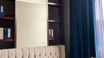 A room with a tufted headboard, a bookshelf with binders, and a window with blue curtains.