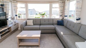 A living room with a grey sectional sofa, a wooden coffee table, a TV on a wooden stand, and large windows with curtains.