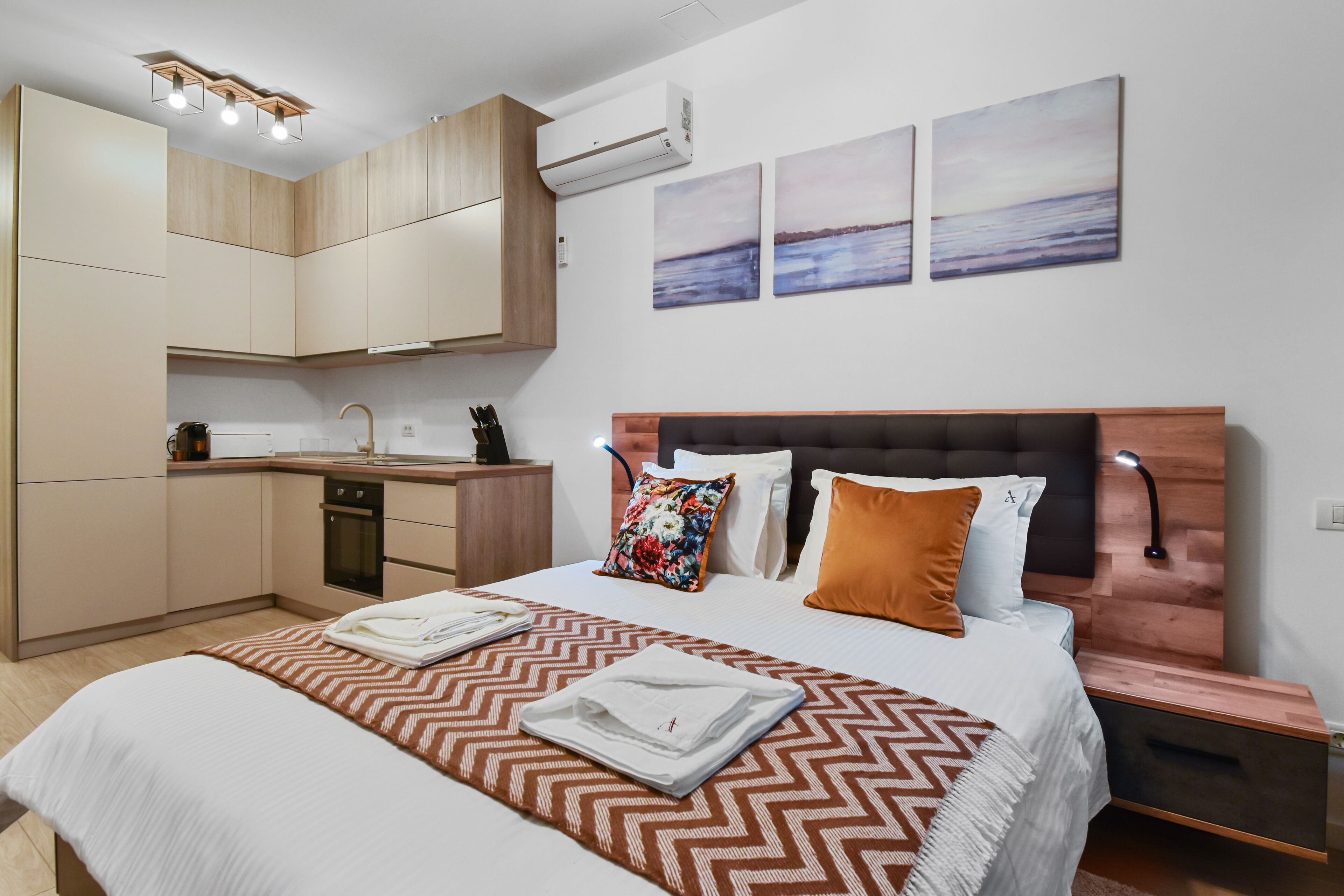Une chambre moderne comprenant un lit, des tables de chevet, une commode, une armoire intégrée, un coin cuisine et des œuvres d’art accrochées au mur.