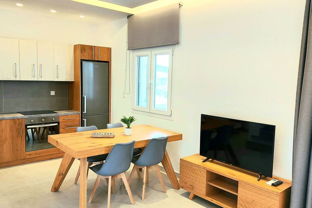 A modern kitchen with wooden cabinets and a built-in stove, a dining area with a wooden table and chairs, and a TV on a wooden stand.